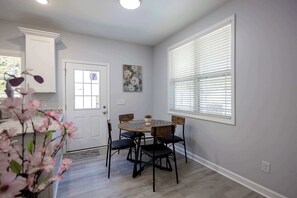 A dining area with a round table and chairs, a white door, a window with blinds, and a painting on the wall.