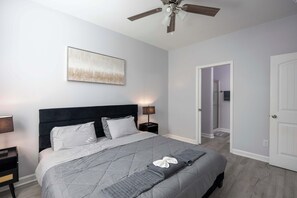 A bedroom with a bed, a ceiling fan, a nightstand, a bedside lamp, and a door leading to another room.