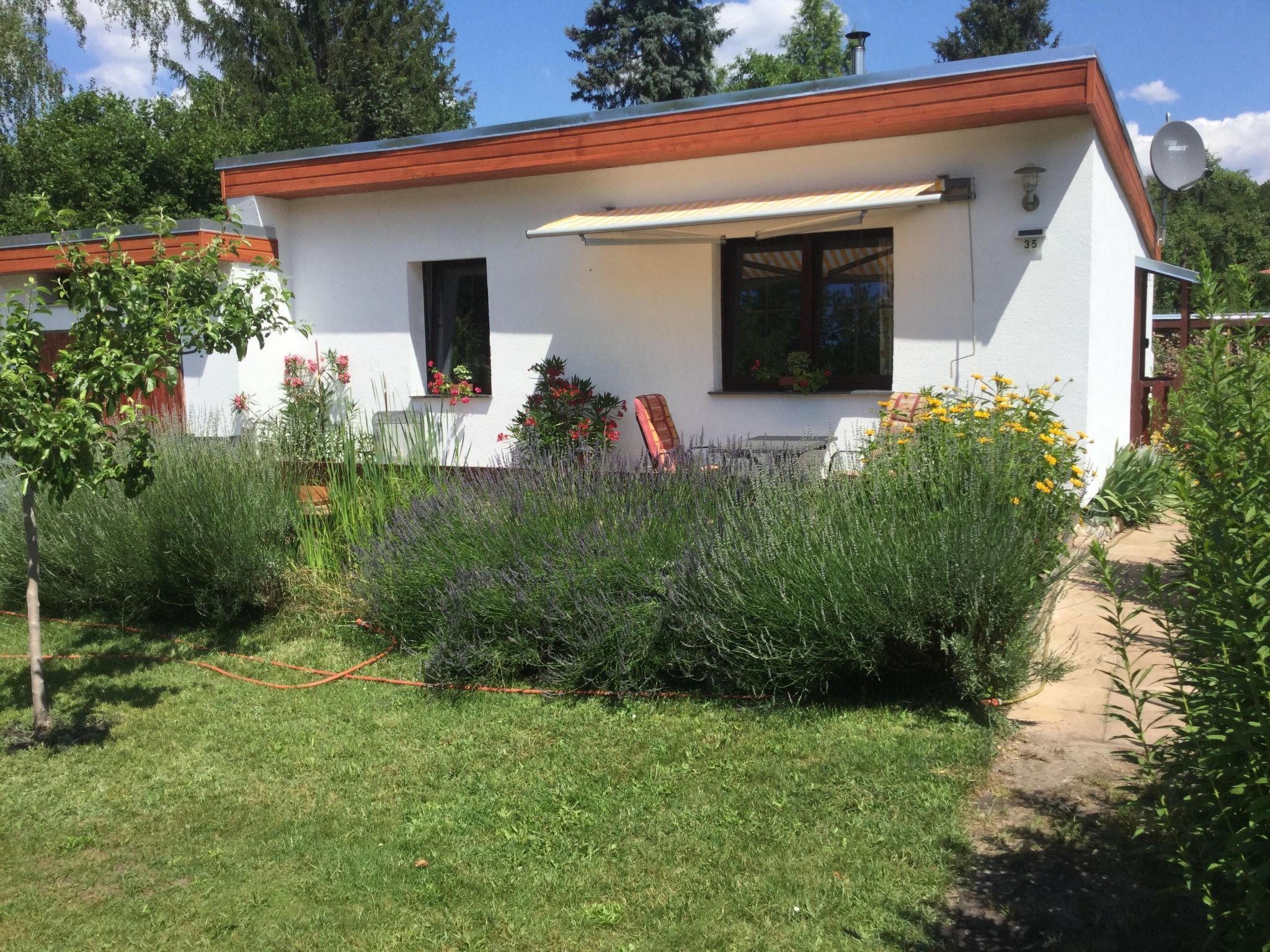 Una casa moderna con fachada blanca y techo rojo, rodeada por un jardín cuidado con diversas plantas y flores.