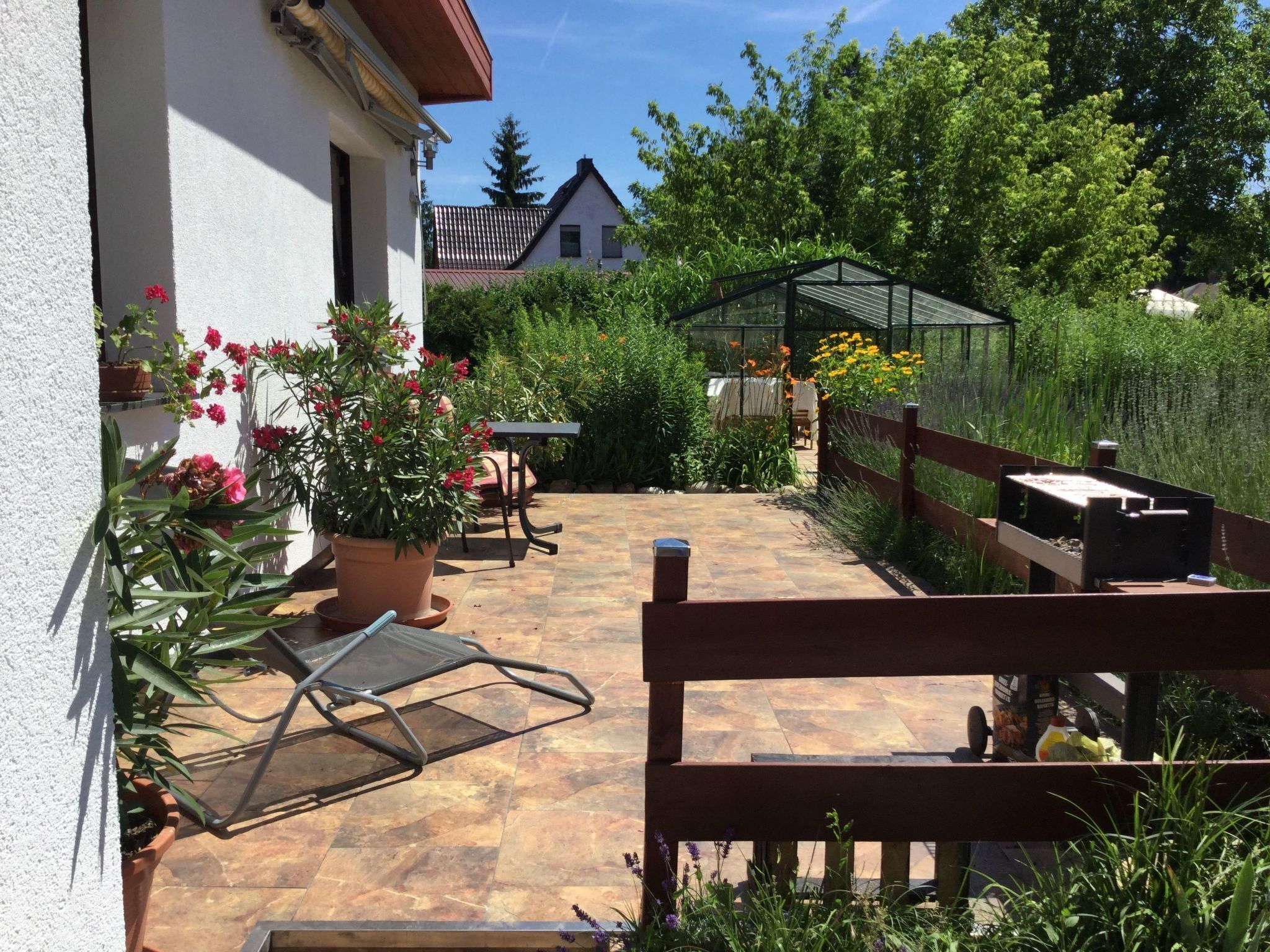 Eine Terrasse mit einer Holzbank, einem Metallstuhl und einem Tisch. Im Hintergrund befinden sich Blumentöpfe und ein Gewächshaus.
