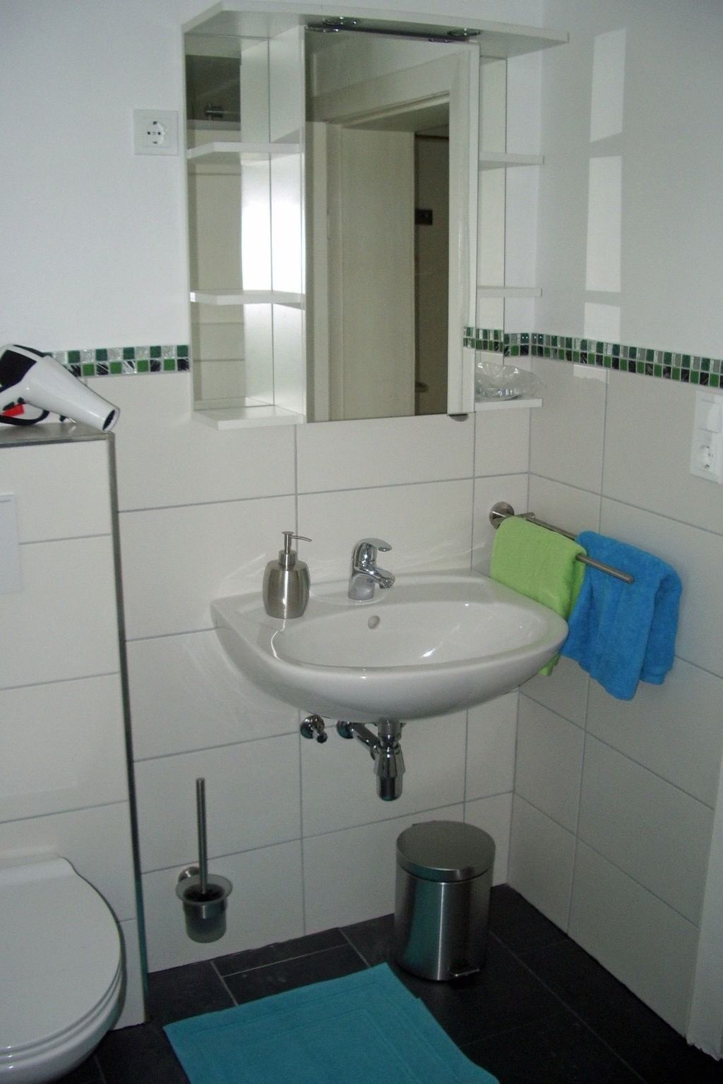 A bathroom with a white sink, a mirror above it, a toilet, a trash bin, and a blue mat on the floor.