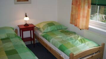 A small, single-room accommodation with two wooden beds, a bedside table with a lamp, a window with curtains, and a radiator.