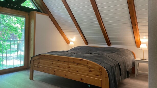 A bedroom with a sloped ceiling, wooden beams, a bed, a nightstand, and a window with a view of greenery. - im Fachwerkstil mit Sauna und Garten by Interhome (Thomasburg)