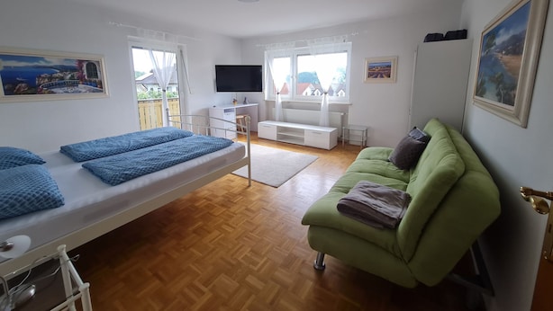Ein Zimmer mit einem Bett, einem grünen Schlafsofa, einem Schreibtisch mit Fernseher und einem Fenster mit Blick auf Häuser.