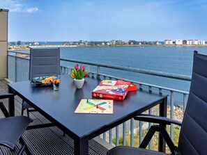 Une table avec un jeu de société, des collations et des fleurs, placée sur un balcon donnant sur une étendue d’eau, avec des bâtiments en arrière-plan.