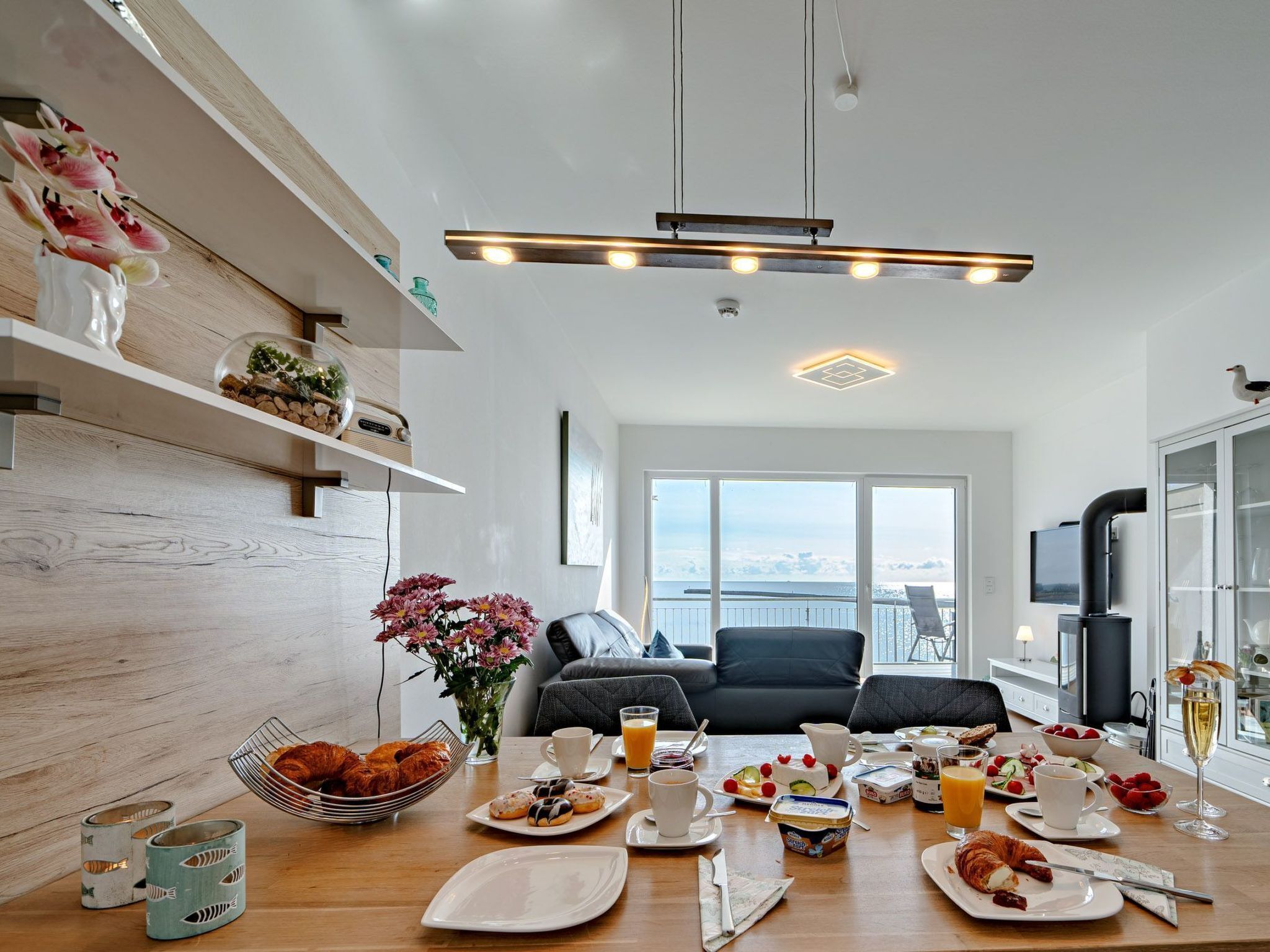 A modern kitchen with a dining table set for breakfast, a large window with a view, and a TV mounted on the wall.