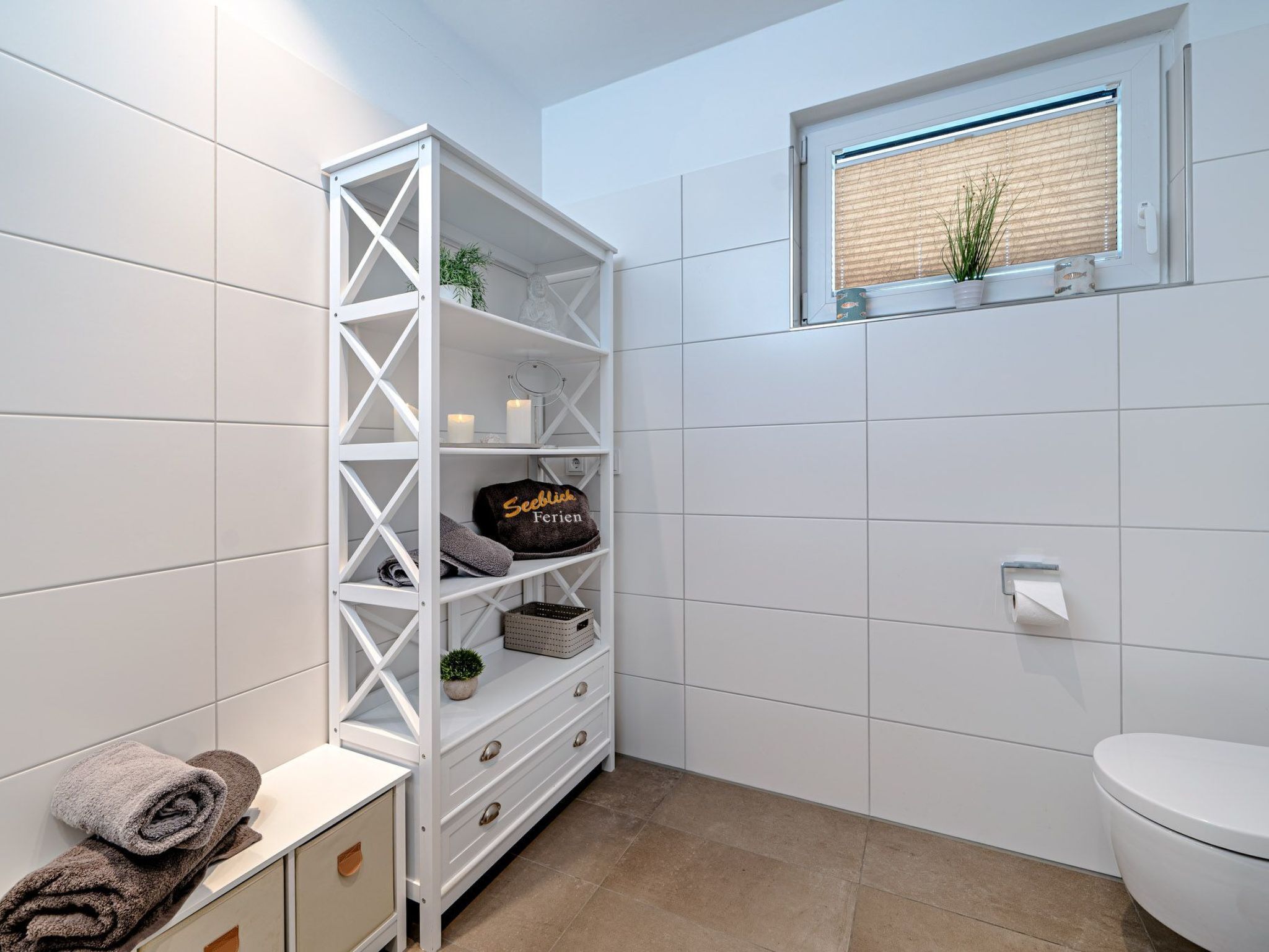 Een badkamer met witte betegelde wanden, een wit toilet, een raam met zonwering, een witte bergkast met planken en handdoeken op een aanrecht.