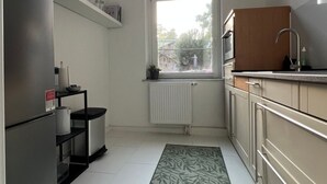 Cocina compacta con refrigerador, estufa y lavamanos. Hay una ventana sobre el lavamanos, un radiador debajo y un tapete estampado en el piso.