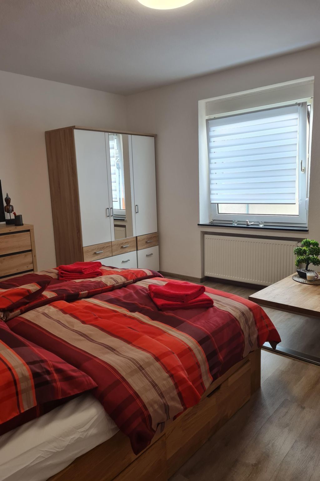 A bedroom with a bed, a wardrobe, a radiator, a window with blinds, and a small table with a plant.