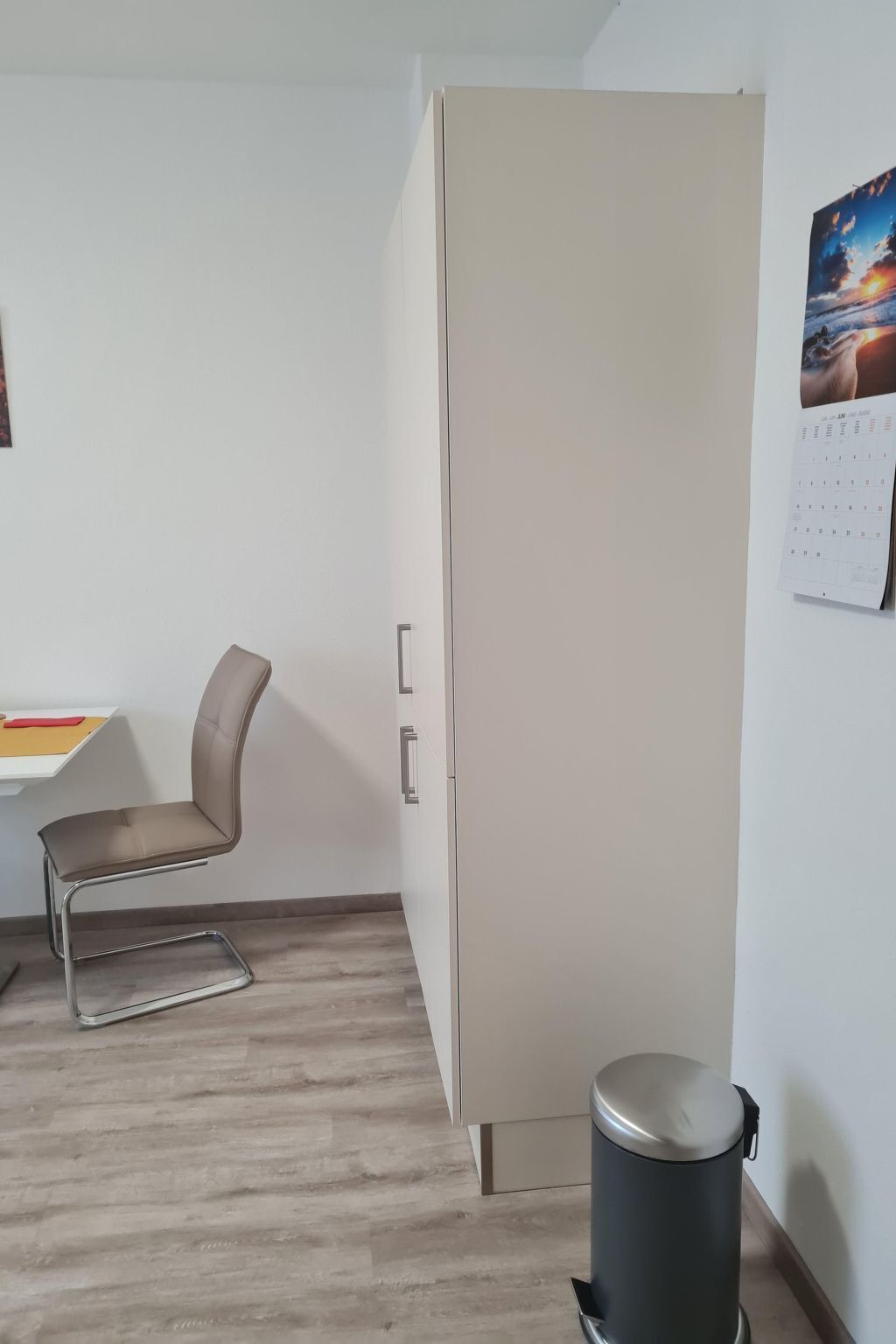 Et værelse med en beige garderobe, et skrivebord med stol, en skraldespand og en kalender på væggen.