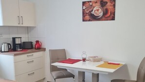 Eine kompakte Küche mit weißen Schränken, einer Kaffeemaschine und einem roten Wasserkocher. Ein Esstisch mit zwei Stühlen steht in der Mitte des Raumes, und ein Bild hängt an der Wand.