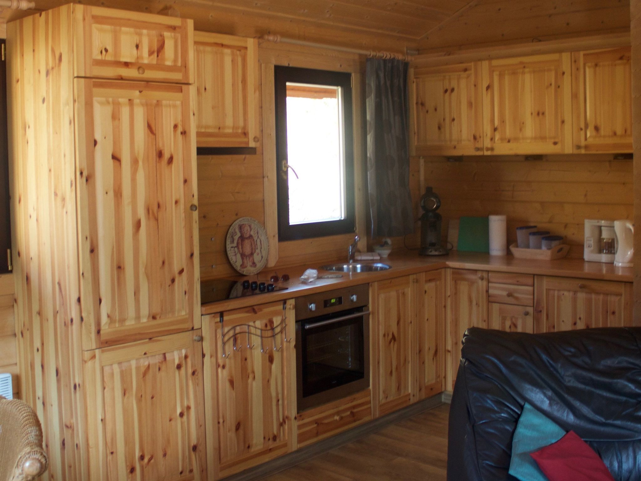 A wooden cabin interior with a kitchen area featuring wooden cabinets, a sink, a stove, and a window with curtains.