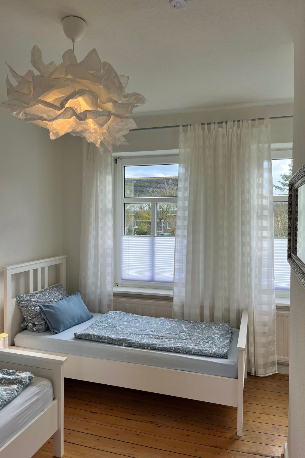 A bedroom with a bed, a window with curtains, a ceiling light fixture, and a view of the outside.