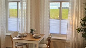 Et spiseområde med et bord dækket til to personer, to stole, en potteplante på et lille bord og en sofa i forgrunden.