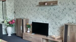 Un soggiorno con televisione su un mobile in legno, due vasi bianchi con fiori e uno scaffale sopra la TV.