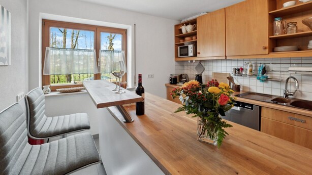 Une cuisine moderne avec des meubles en bois, un coin repas avec un banc et une vue sur les arbres par la fenêtre.