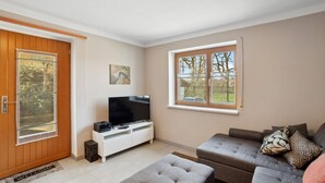 Un salon comprenant un téléviseur, un canapé, un pouf et une porte donnant sur une vue sur les arbres.