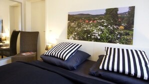 Una camera da letto con un letto, due cuscini, una sedia, un comodino e un quadro appeso al muro raffigurante un giardino.