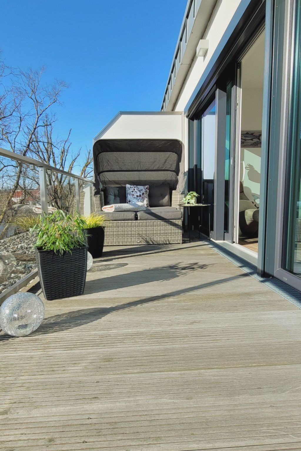 A modern outdoor seating area with a sofa and cushions, potted plants, and a glass door leading to a room.