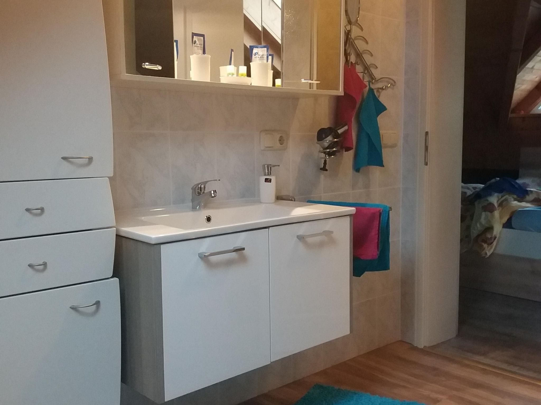 A modern bathroom with a white sink, mirror, and cabinets. There are towels hanging on hooks and a shower area visible in the background.