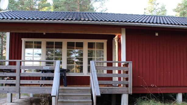 Punaisella maalilla maalattu puinen mökki, jossa on kuisti, pöytä ja tuolit sekä portaat, jotka johtavat sisäänkäynnille.
