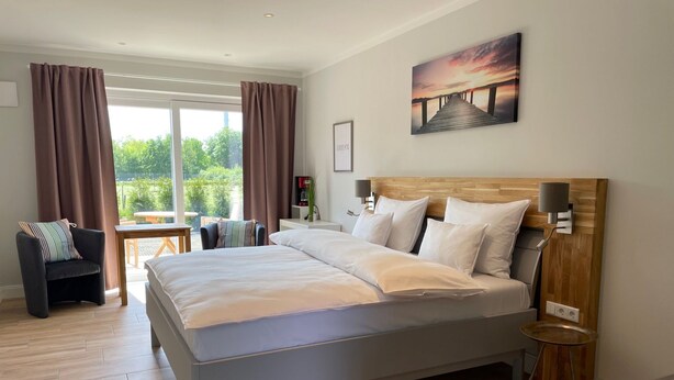 Una camera da letto moderna con un letto grande, una testiera in legno, un comodino, una scrivania e una sedia. Si vede una finestra con tende, un quadro appeso al muro e una vista sul verde all'esterno.