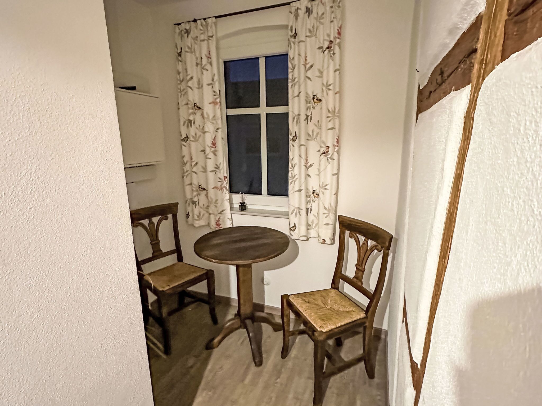Una habitación con una mesa redonda de madera y dos sillas de madera, una ventana con cortinas florales y una pared blanca.