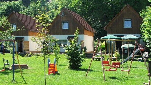 Un terrain de jeux avec des balançoires et des toboggans, entouré de deux maisons en bois aux toits rouges et aux murs blancs.
