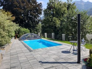 A swimming pool with a glass enclosure, surrounded by a paved patio and a garden with trees and a fence. - Apartment Anna by Interhome (Flattach)