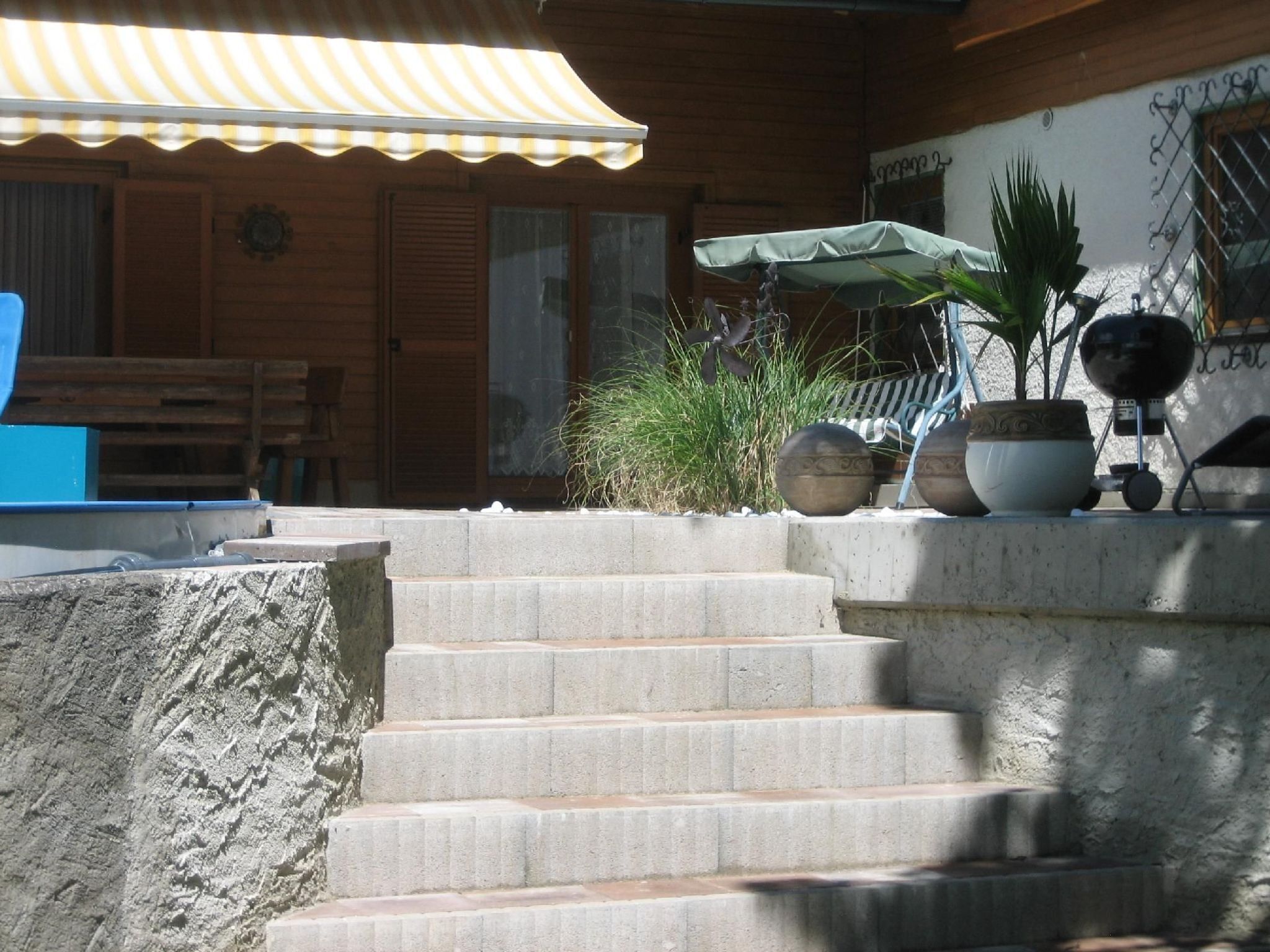 A set of concrete steps leading to a wooden door, a concrete planter with a potted plant, and a blue object on the left.