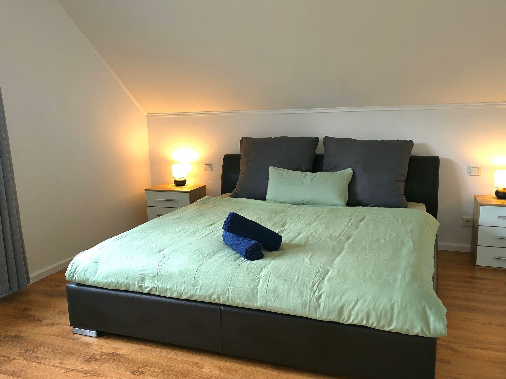 A neatly made bed with a green bedspread and two pillows, a bedside table with a lamp, and a wooden floor.