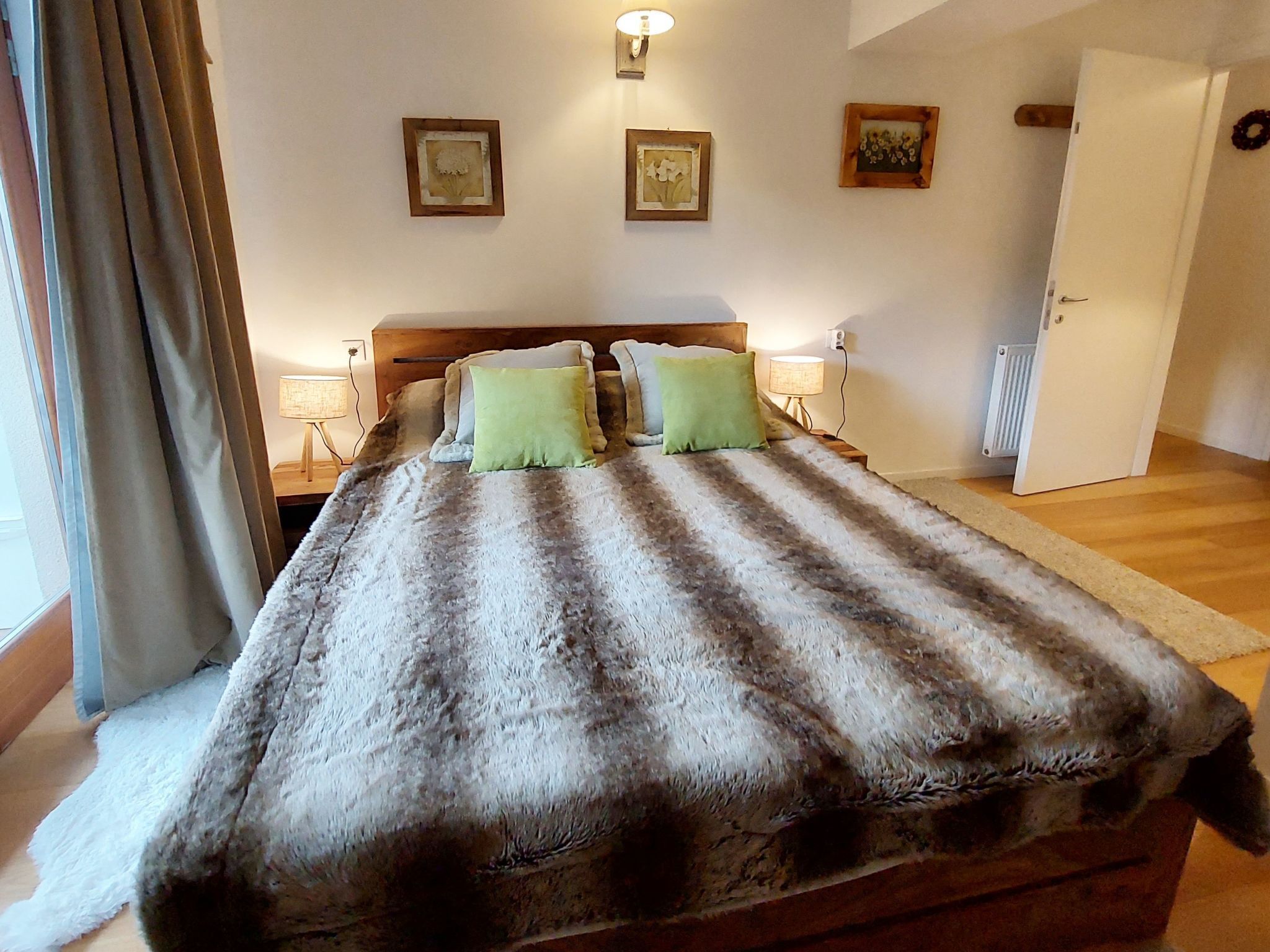 A bedroom with a large bed, wooden headboard, and two bedside lamps. There are three framed pictures on the wall and a door to the right.