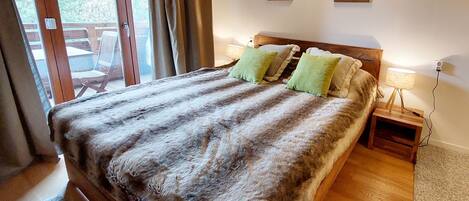 Een slaapkamer met een groot bed, een houten hoofdeinde, twee nachtkastjes, een lamp en uitzicht op het omliggende landschap door een schuifdeur.
