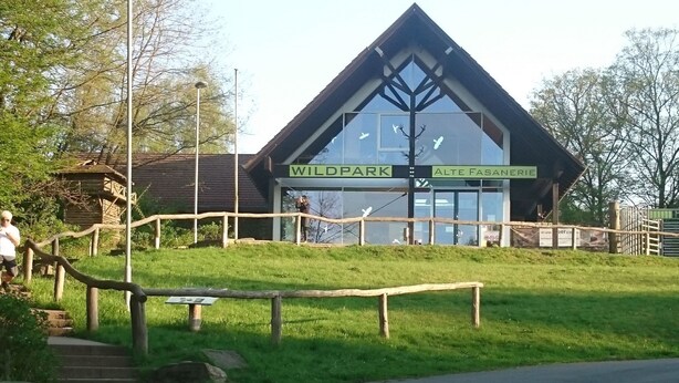 Ein Gebäude mit der Aufschrift "Wildpark Alte Fasane", umgeben von einem Holzzaun und mit einem befestigten Weg, der dorthin führt.