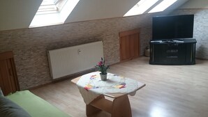 Ein Loft-Zimmer mit schrägem Dach, Holzboden, einem grünen Sofa, einem kleinen Holztisch mit Blumenmuster und einem Topf mit Pflanze sowie einer schwarzen Unterhaltungsanlage mit Flachbildfernseher.