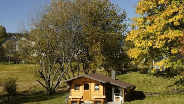 Una cabaña de madera con un porche pequeño, ubicada junto a un estanque que refleja los árboles circundantes.