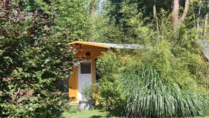 Una piccola costruzione in legno giallo con una porta bianca, immersa nel verde e circondata da alberi.