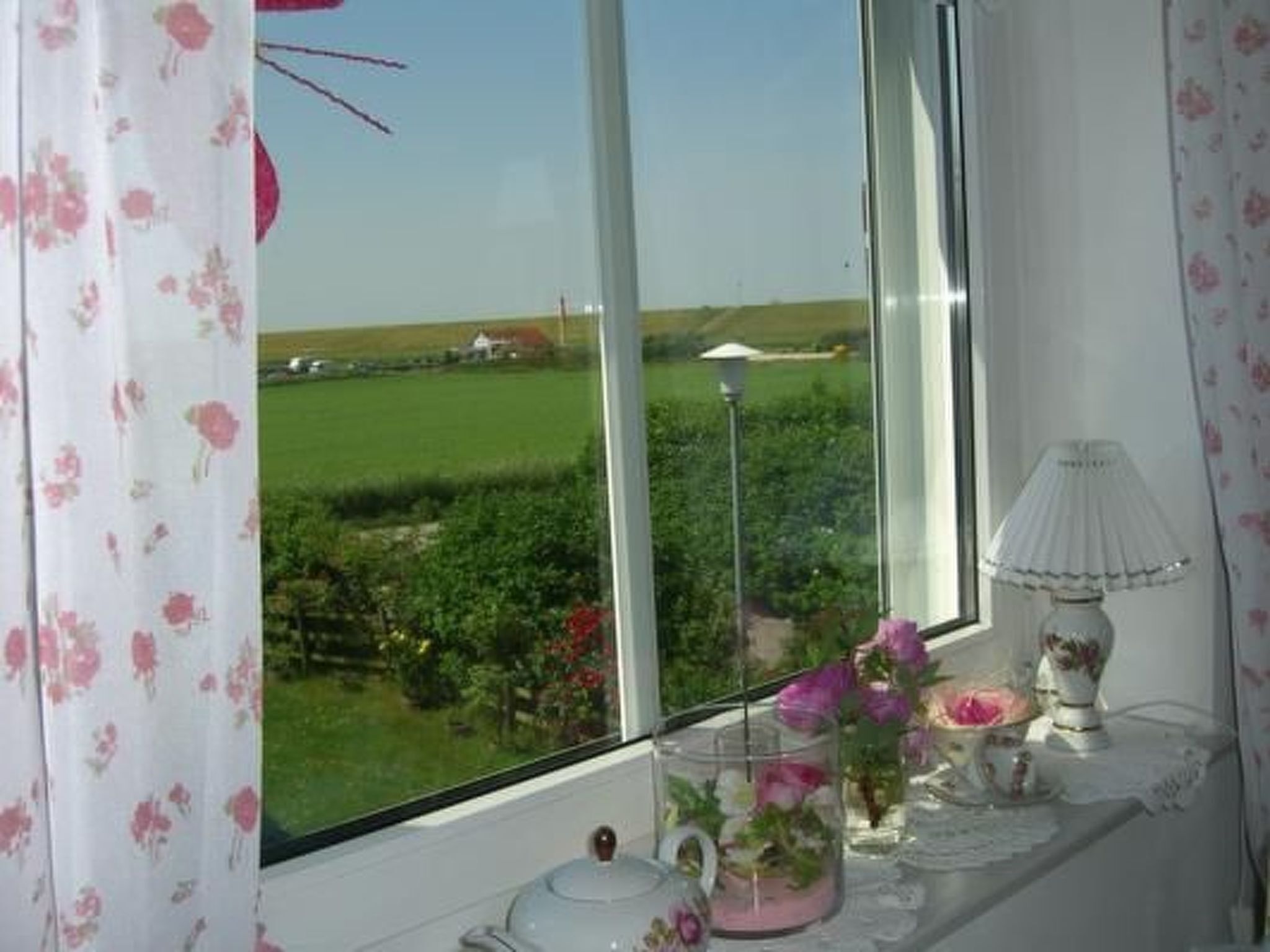 Una habitación con un ventanal grande que da a un campo verde y un edificio blanco. Hay una mesa con un hervidor, una lámpara y un florero con flores.