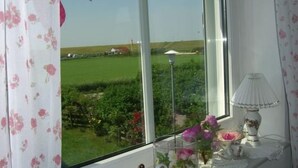 Una habitación con un ventanal grande que da a un campo verde y un edificio blanco. Hay una mesa con un hervidor, una lámpara y un florero con flores.