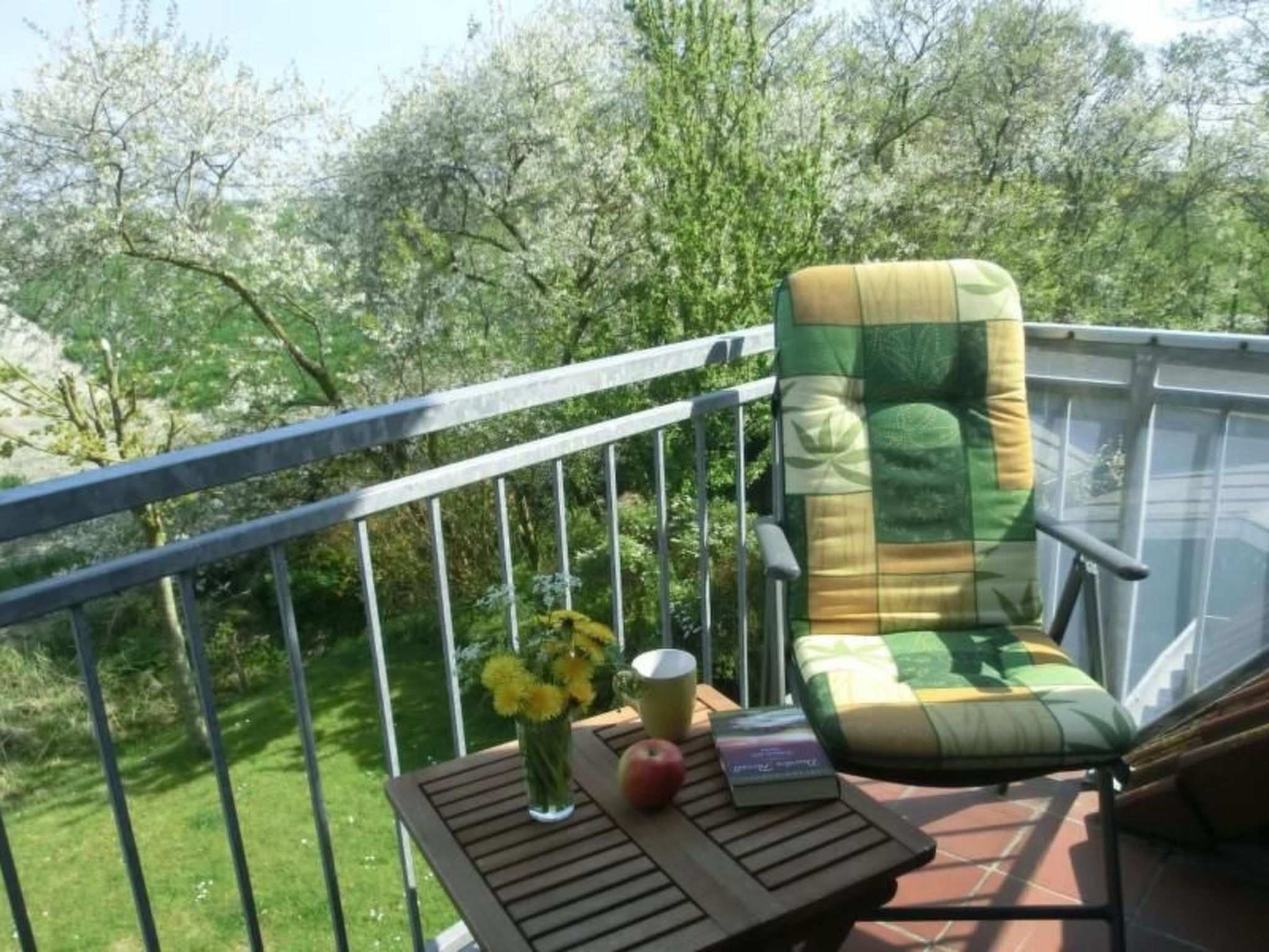 A balcony with a cushioned chair, a small table with a book, a vase with flowers, and a glass of drink.