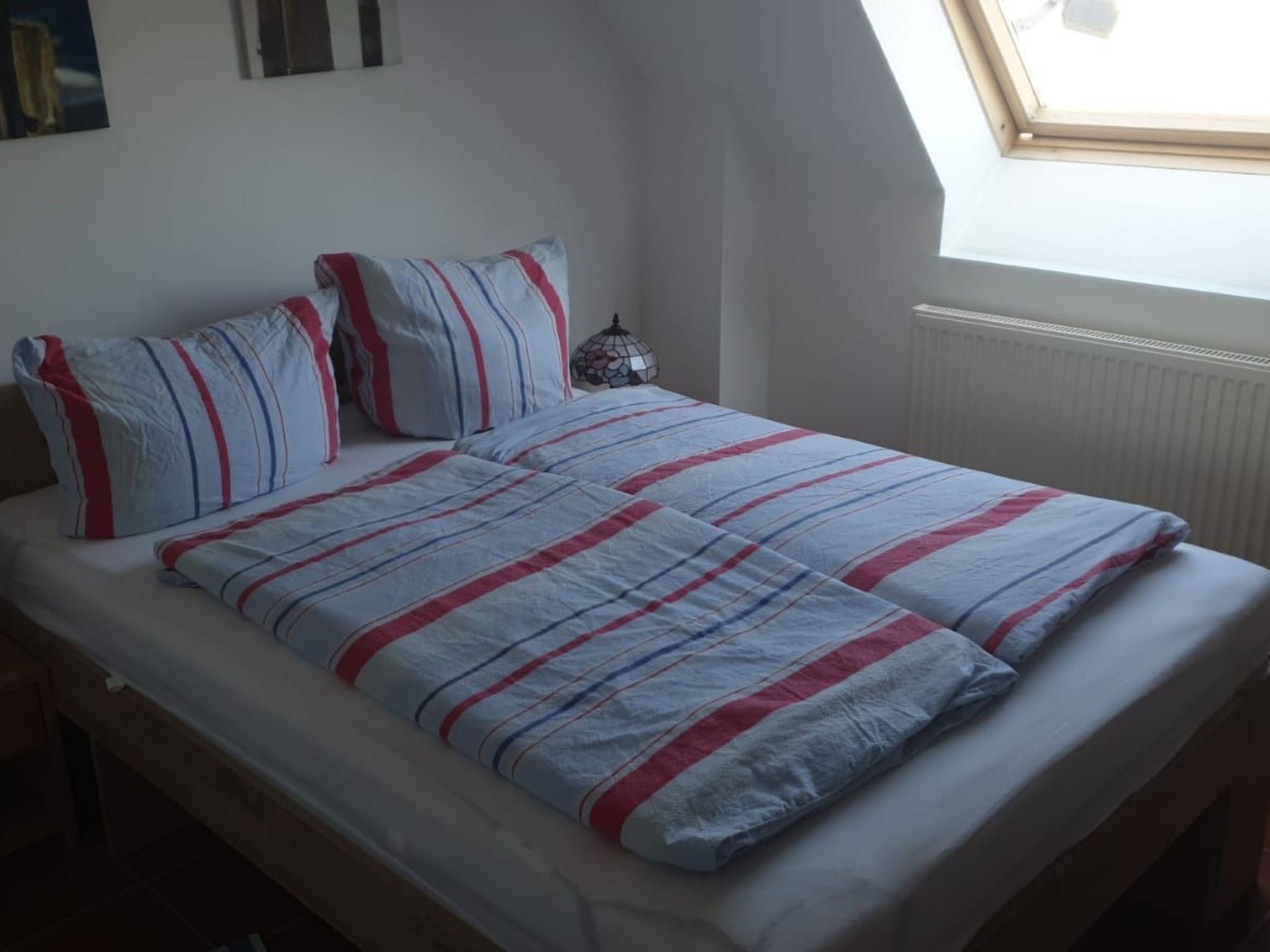 Un dormitorio con cama, ropa de cama a rayas, una mesita de noche, un reloj de pared y una ventana con vistas.