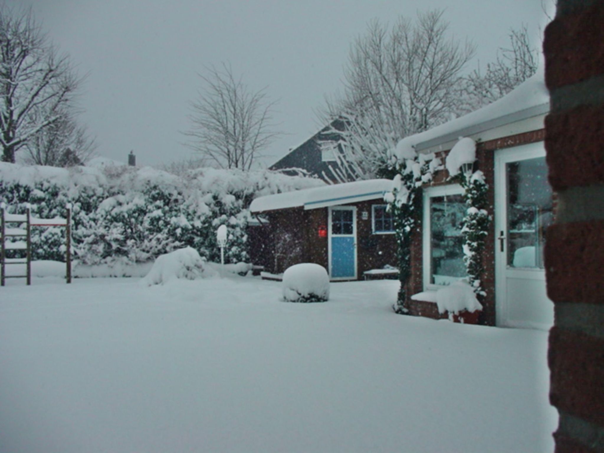 A snow-covered residential area with a brick house, a blue door, and a visible garage.
