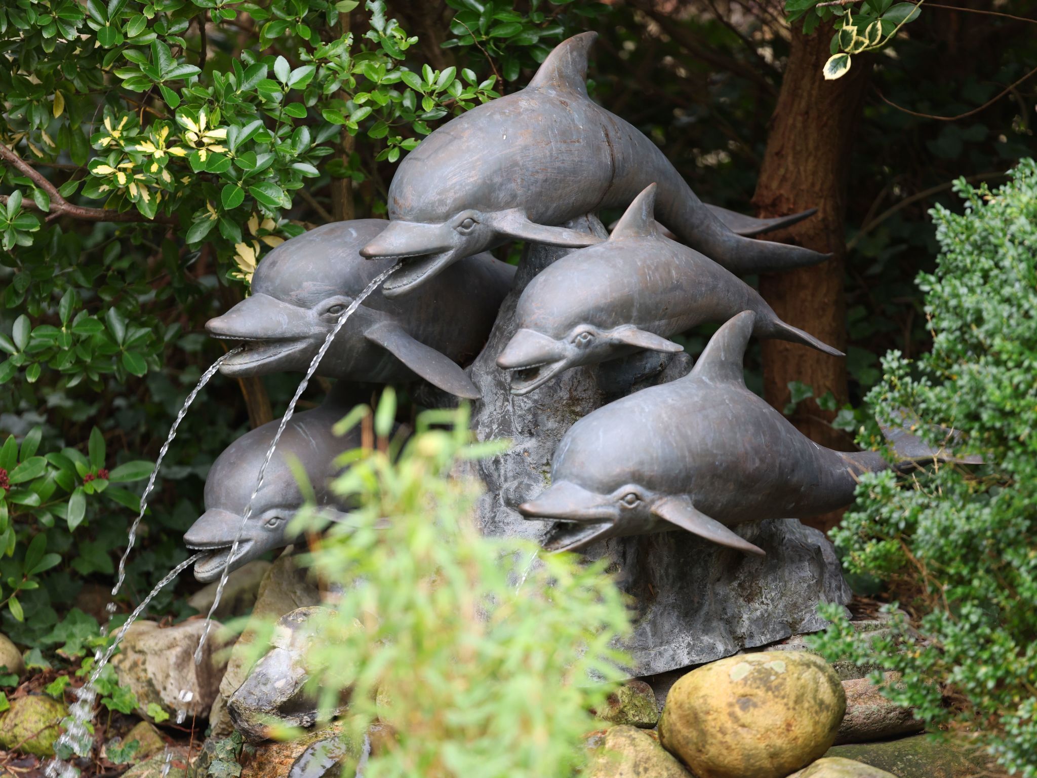 Escultura de golfinhos com água jorrando das bocas, em meio a rochas e vegetação.