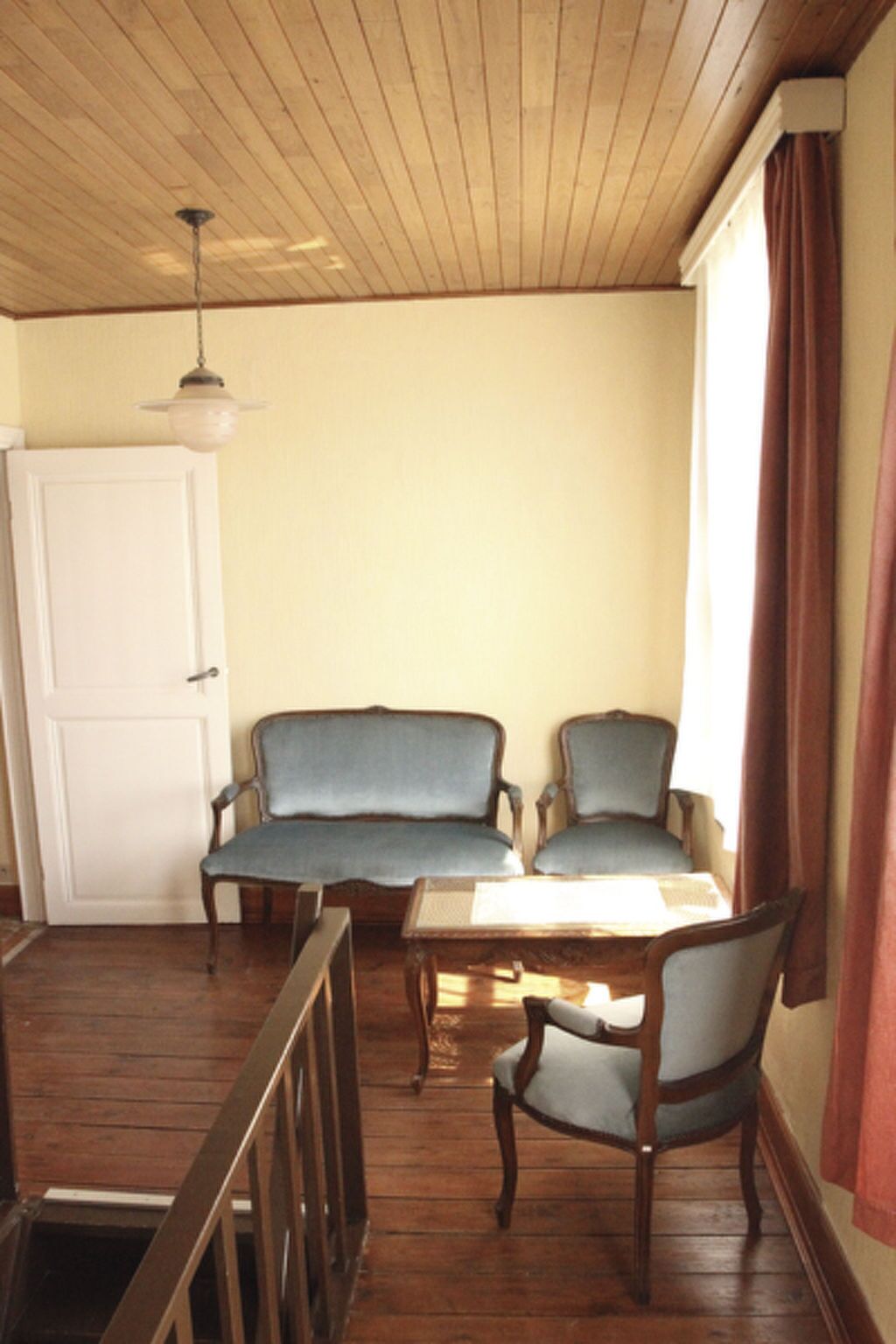 Une pièce avec un plancher en bois, deux fauteuils rembourrés, une table en bois, une porte blanche et une fenêtre avec des rideaux.