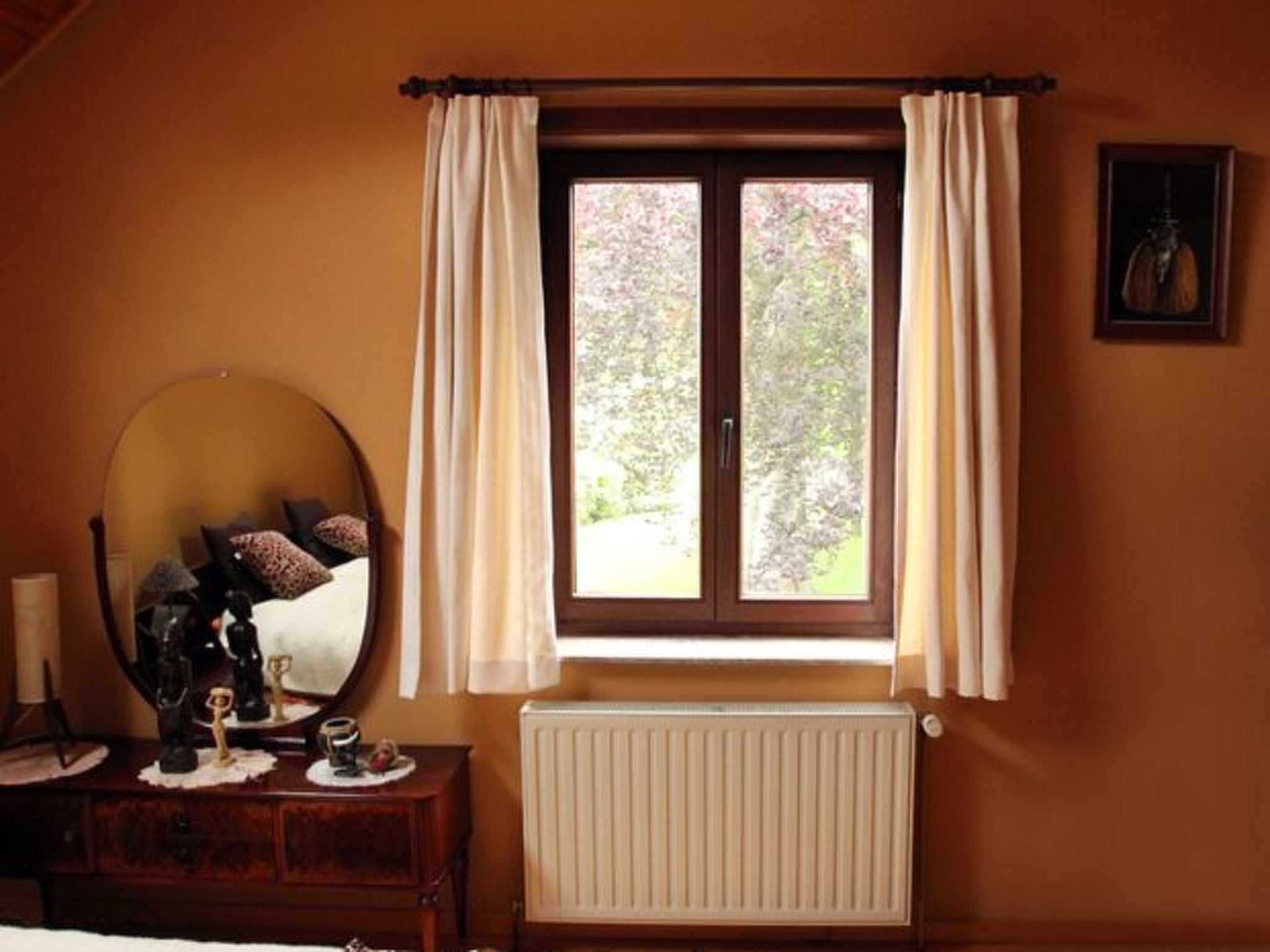 Ein Schlafzimmer mit einem großen Spiegel, einer Kommode mit Dekorationen, einem Fenster mit Vorhängen und einem Heizkörper.
