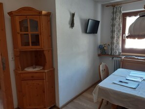 Ein Zimmer mit einem Holzkabinett, einem an der Wand befestigten Fernseher, einem Tisch mit Papieren und einem Fenster mit Vorhängen.