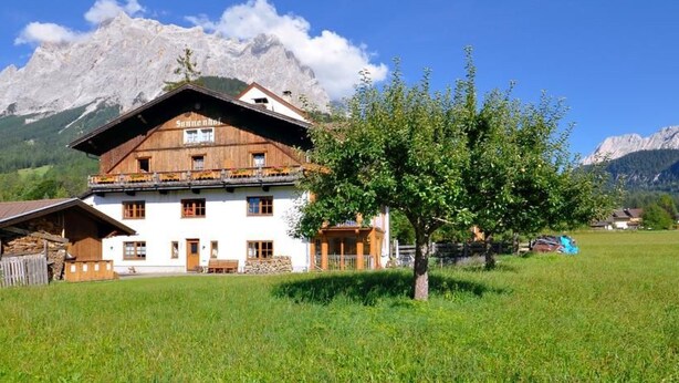 En traditionel schweizisk chalet med træfacade, mange vinduer og en balkon. Den ligger i en græsplæne med et bjerglandskab i baggrunden.