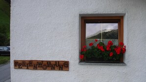 En skylt med texten ”FERIENWUNGS” på en vit vägg, ett fönster med röda blommor och utsikt över ett berg.