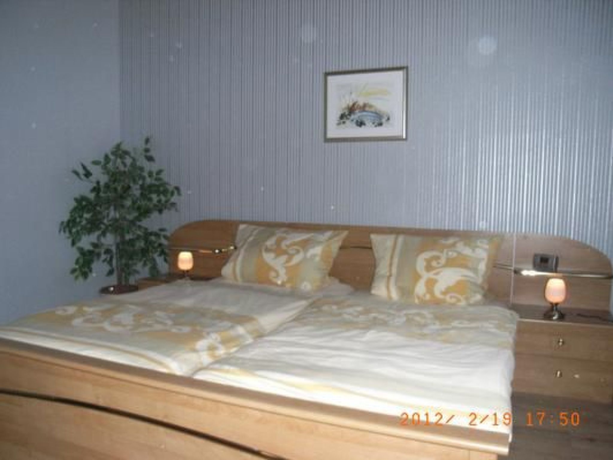 A bedroom with a bed, bedside tables, a lamp, a framed picture on the wall, and a potted plant.