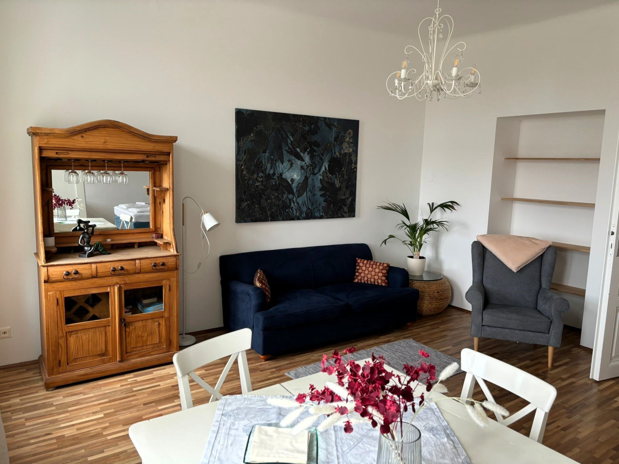 Un salon avec un meuble en bois, un canapé bleu foncé, une table blanche ornée d’un vase de fleurs et un grand tableau abstrait accroché au mur.
